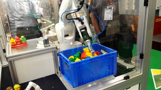 Un robot en una celda introduce la mano en una caja azul que contiene objetos de colores